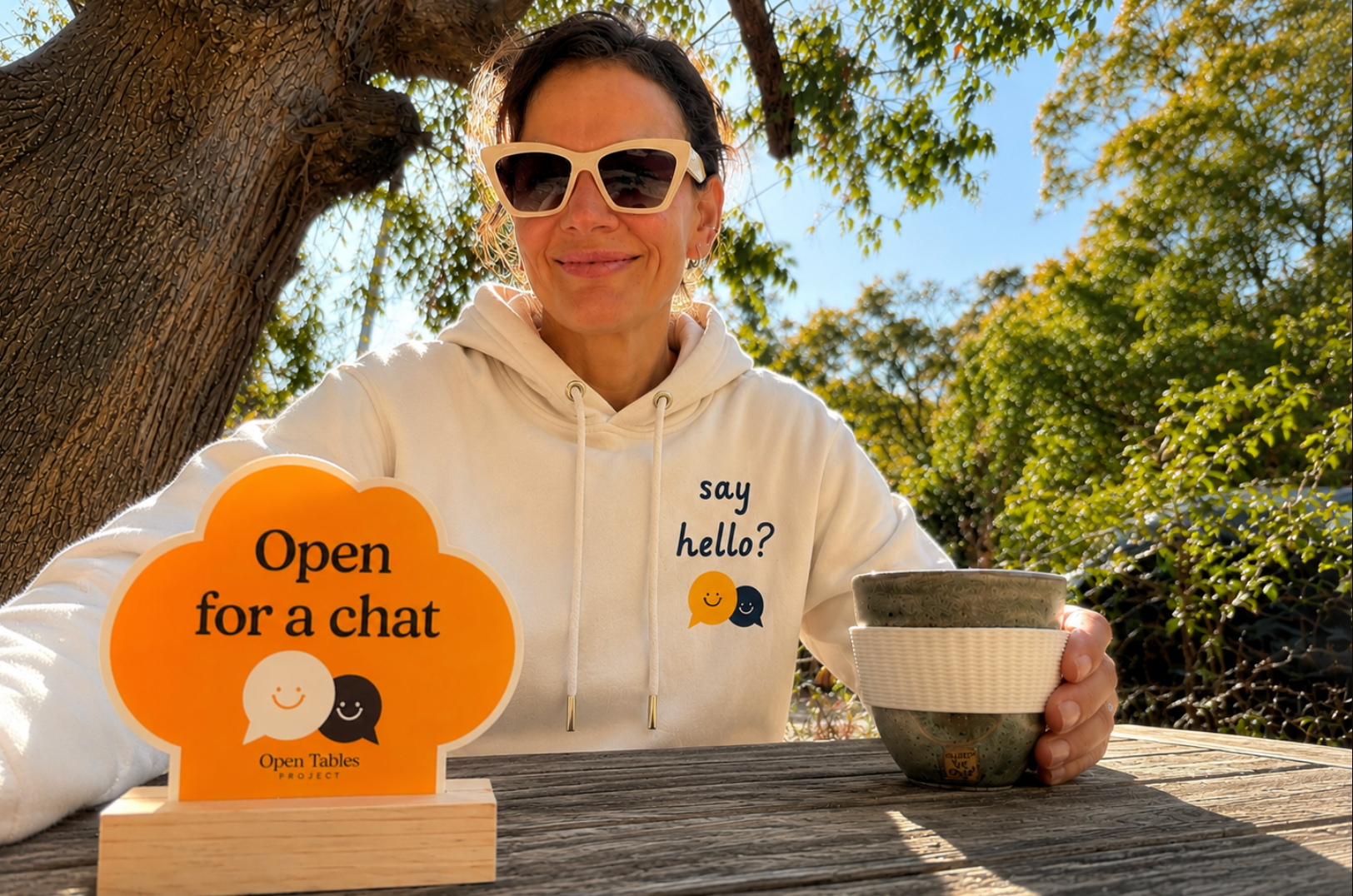 Founder of Open Tables Project wearing the Say Hello hoodie while sitting with an Open for a Chat marker and coffee outdoors.